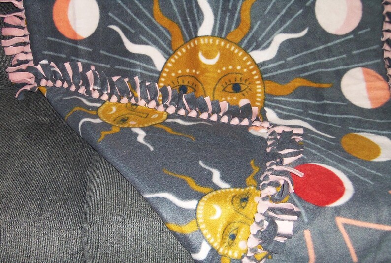 May include: A gray fleece blanket with a repeating pattern of suns and moons. The suns are yellow with a crescent moon inside and have a stylized face with eyes and a nose. The moons are white with pink accents. The blanket has a pink and gray knotted fringe border.