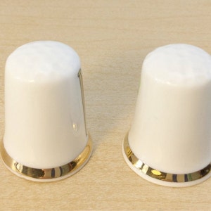 May include: Two white ceramic thimbles with a gold trim around the top edge.