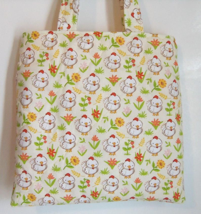 May include: A quilted tote bag made of cream-colored fabric featuring a repeating pattern of white chickens with red combs, yellow sunflowers, and green grass stalks. The bag includes two matching fabric handles at the top for carrying.
