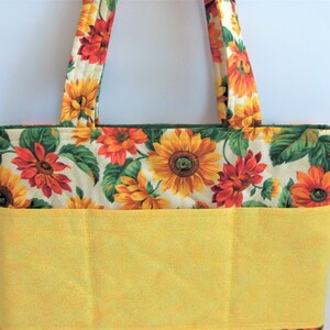 May include: A yellow and green tote bag with a sunflower print. The bag has a large pocket on the front with two smaller pockets on either side.
