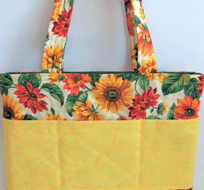 May include: A yellow and green tote bag with a sunflower print. The bag has a large pocket on the front with two smaller pockets on either side.