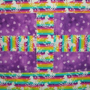 May include: A quilted fabric item featuring alternating horizontal bands of a rainbow-striped pattern with white unicorn silhouettes and a solid purple fabric panel decorated with a repeating pattern of small, light-colored heart shapes.