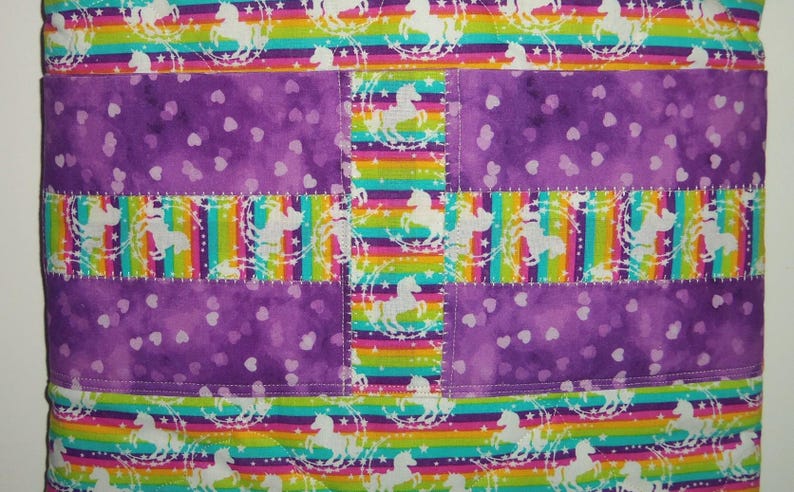 May include: A quilted fabric item featuring alternating horizontal bands of a rainbow-striped pattern with white unicorn silhouettes and a solid purple fabric panel decorated with a repeating pattern of small, light-colored heart shapes.