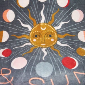 May include: A gray fleece blanket with a sun and moon design. The sun is yellow with a face and the moon is white with a crescent shape. The blanket has a pink fringe around the edges.