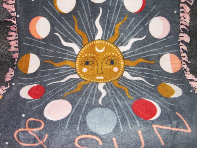 May include: A gray fleece blanket with a sun and moon design. The sun is yellow with a face and the moon is white with a crescent shape. The blanket has a pink fringe around the edges.