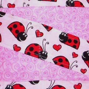 May include: A fabric textile featuring a repeating pattern of red and black ladybugs interspersed with small red hearts on a light pink background, layered with sections of a light pink fabric displaying a tonal swirling spiral pattern.