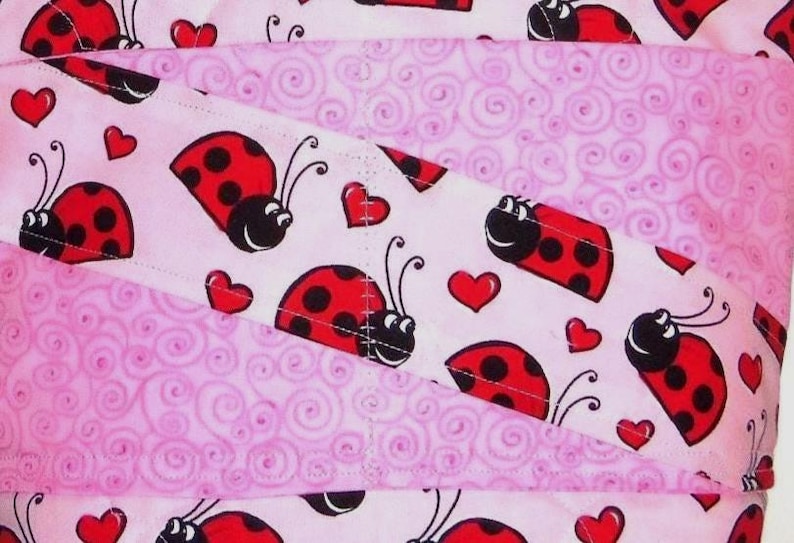 May include: A fabric textile featuring a repeating pattern of red and black ladybugs interspersed with small red hearts on a light pink background, layered with sections of a light pink fabric displaying a tonal swirling spiral pattern.