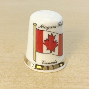 May include: White ceramic thimble with a gold trim. It features a Canadian flag with the words "Niagara Falls" and "Canada" printed on it.