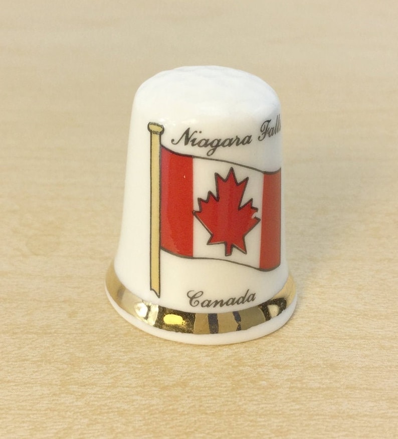 May include: White ceramic thimble with a gold trim. It features a Canadian flag with the words "Niagara Falls" and "Canada" printed on it.