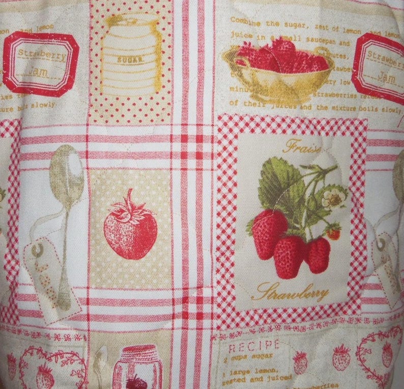 May include: A quilted fabric featuring a grid pattern with red and white gingham borders. The design includes illustrations of strawberries, a bowl of berries, a sugar jar, and a spoon, alongside printed text about strawberry jam recipes.