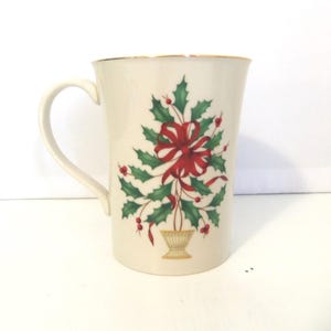 May include: A cream-colored ceramic mug featuring a central graphic of a stylized holly plant arranged in a small, tan-colored urn. The design includes green holly leaves, red berries, and a prominent red ribbon bow. The mug has a gold-toned rim.