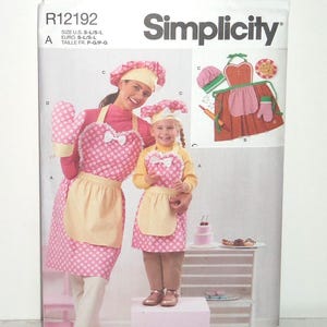 May include: Simplicity sewing pattern featuring a mother and child wearing matching pink polka dot aprons, oven mitts, and chef hats. The pattern includes a diagram of the apron design and the words "easy-to-sew".