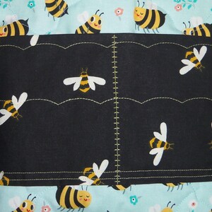 K&ouml;nnte beinhalten: Eine gesteppte Stofftasche mit hellblauem Hintergrund und einem sich wiederholenden Muster aus gelben und schwarzen Bienen sowie kleinen Blumen. Ein schwarzes horizontales Taschenpanel ist mittig aufgen&auml;ht, verziert mit Bienenmuster.