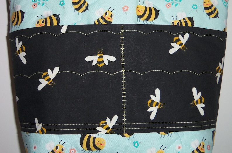 K&ouml;nnte beinhalten: Eine gesteppte Stofftasche mit hellblauem Hintergrund und einem sich wiederholenden Muster aus gelben und schwarzen Bienen sowie kleinen Blumen. Ein schwarzes horizontales Taschenpanel ist mittig aufgen&auml;ht, verziert mit Bienenmuster.