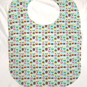 May include: A baby bib featuring a light blue background adorned with a pattern of colourful cupcakes. The bib has a large neck opening and a rectangular shape with rounded edges. The cupcakes are in various colours.