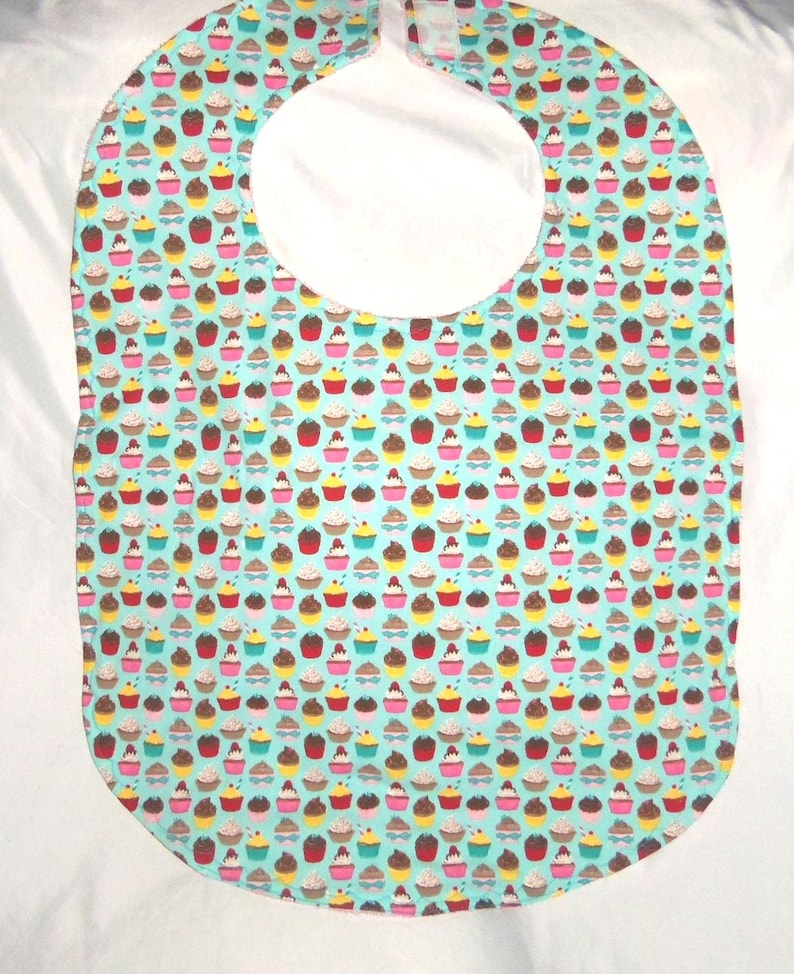 May include: A baby bib featuring a light blue background adorned with a pattern of colourful cupcakes. The bib has a large neck opening and a rectangular shape with rounded edges. The cupcakes are in various colours.
