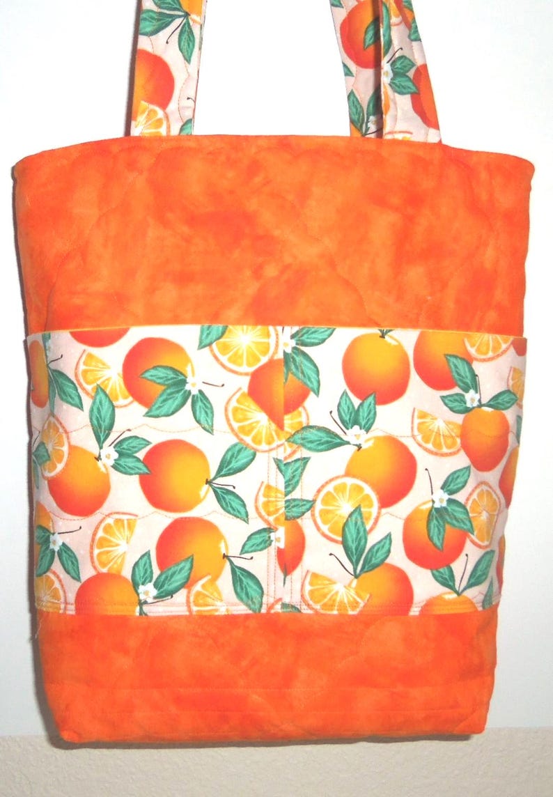 May include: Orange tote bag with a vibrant orange fabric and a band of white fabric featuring orange fruit illustrations. The handles are made of the same orange fruit pattern fabric. The bag is designed for carrying items.