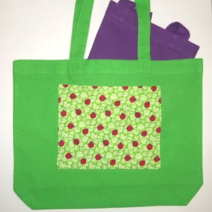K&ouml;nnte beinhalten: Eine leuchtend gr&uuml;ne Tragetasche mit einer rechteckigen Vordertasche, die ein wiederkehrendes Muster aus roten Marienk&auml;fern auf hellgr&uuml;nem Blattgrund zeigt. Eine zweite, einfarbige violette Tragetasche ist teilweise darin sichtbar.