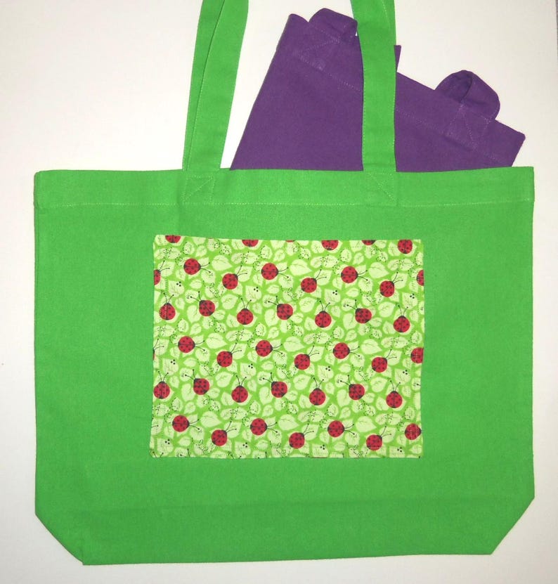 K&ouml;nnte beinhalten: Eine leuchtend gr&uuml;ne Tragetasche mit einer rechteckigen Vordertasche, die ein wiederkehrendes Muster aus roten Marienk&auml;fern auf hellgr&uuml;nem Blattgrund zeigt. Eine zweite, einfarbige violette Tragetasche ist teilweise darin sichtbar.