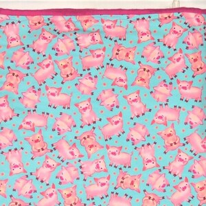 May include: A rectangular, light blue fabric with a repeating pattern of pink cartoon pigs in various poses. The fabric has a pink border. The pigs are in different positions, some upside down, with small orange dots scattered throughout.