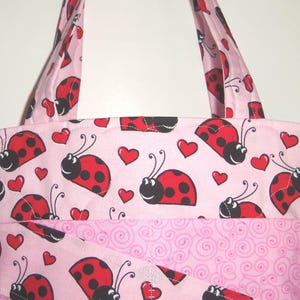 May include: A quilted tote bag featuring a light pink background with a repeating pattern of red ladybugs and small red hearts. The bag includes two fabric handles and a lower section with a light pink swirl pattern and a diagonal fabric overlay.