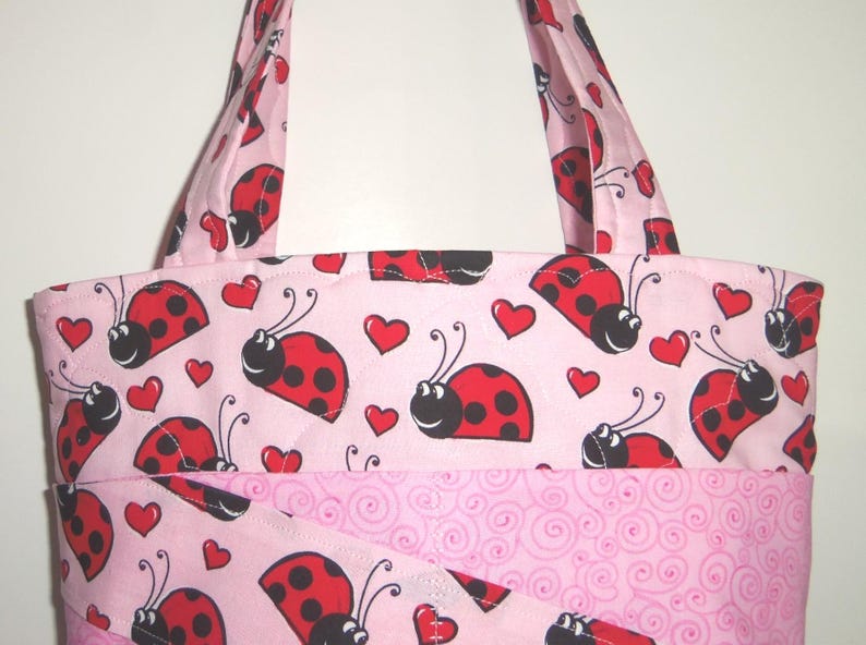 May include: A quilted tote bag featuring a light pink background with a repeating pattern of red ladybugs and small red hearts. The bag includes two fabric handles and a lower section with a light pink swirl pattern and a diagonal fabric overlay.