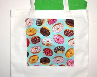 May include: A white tote bag featuring a central square panel with a light blue background printed with various colorful donuts with icing and sprinkles. A bright green fabric item is tucked inside the bag.