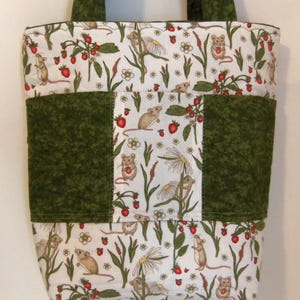 May include: A quilted tote bag featuring a white fabric with a repeating pattern of brown mice, red strawberries, and white daisy flowers. The bag includes solid dark green fabric panels on the sides and center, with matching dark green fabric handles.