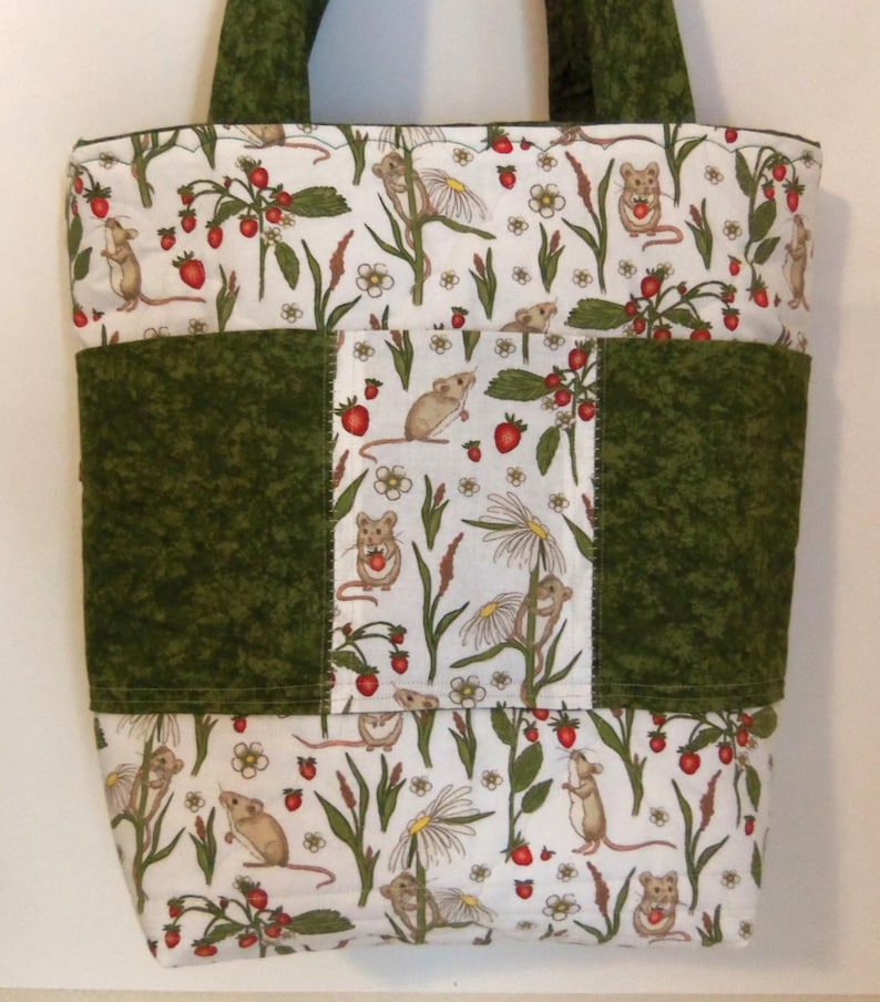 May include: A quilted tote bag featuring a white fabric with a repeating pattern of brown mice, red strawberries, and white daisy flowers. The bag includes solid dark green fabric panels on the sides and center, with matching dark green fabric handles.
