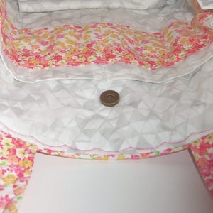 May include: A quilted tote bag interior featuring a white fabric with a geometric triangle pattern and a scalloped pocket edge. The pocket is lined with a vibrant floral print in shades of pink, orange, and yellow. A single metal magnetic snap closure.