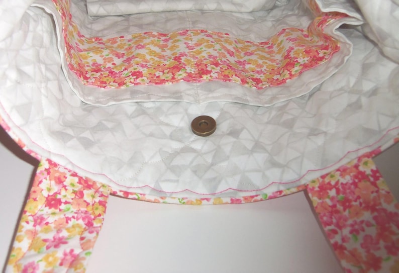 May include: A quilted tote bag interior featuring a white fabric with a geometric triangle pattern and a scalloped pocket edge. The pocket is lined with a vibrant floral print in shades of pink, orange, and yellow. A single metal magnetic snap closure.
