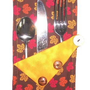 May include: A brown fabric napkin with a pattern of orange, red, and yellow leaves. The napkin has a yellow fabric pocket with two brown buttons and holds a fork, knife, and spoon.
