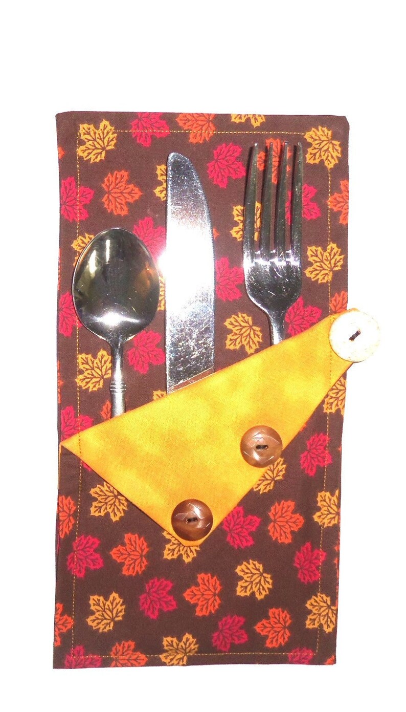 May include: A brown fabric napkin with a pattern of orange, red, and yellow leaves. The napkin has a yellow fabric pocket with two brown buttons and holds a fork, knife, and spoon.