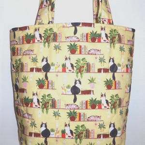 May include: A quilted tote bag made of yellow fabric featuring a repeating pattern of cats sitting on shelves alongside potted green plants and colorful books. The bag includes two matching fabric shoulder straps and a vertical rectangular shape.