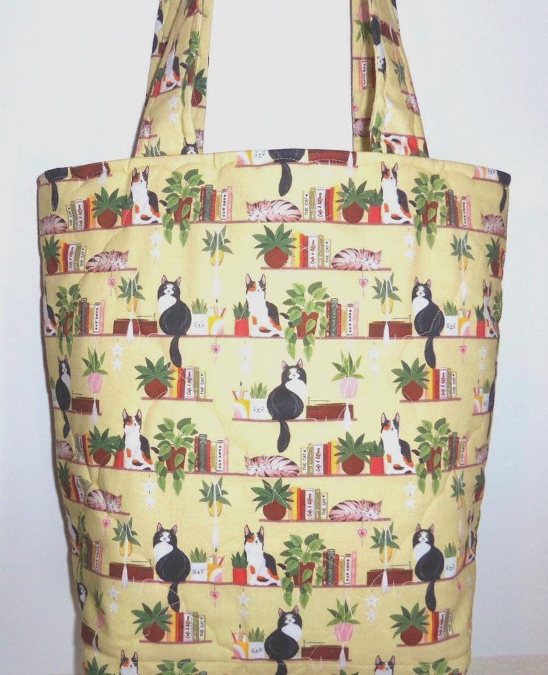 May include: A quilted tote bag made of yellow fabric featuring a repeating pattern of cats sitting on shelves alongside potted green plants and colorful books. The bag includes two matching fabric shoulder straps and a vertical rectangular shape.