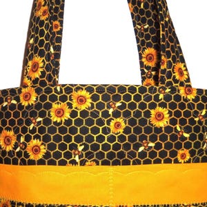 May include: A quilted tote bag featuring a black fabric background with a yellow honeycomb pattern, yellow sunflowers, and small bees. The bag includes two matching fabric handles and a solid yellow horizontal band with a scalloped top edge.