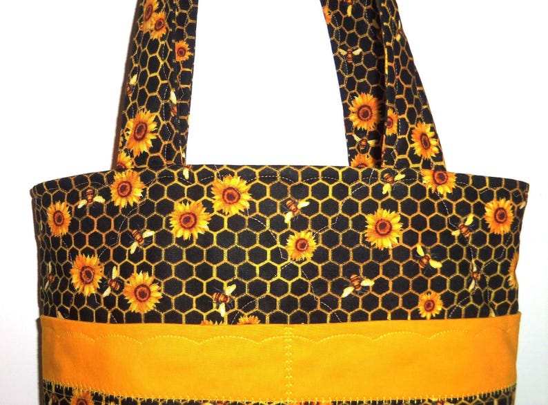 May include: A quilted tote bag featuring a black fabric background with a yellow honeycomb pattern, yellow sunflowers, and small bees. The bag includes two matching fabric handles and a solid yellow horizontal band with a scalloped top edge.