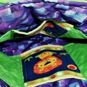 May include: A quilted fabric bag interior featuring a vibrant purple and blue checkered pattern lining. The bag includes bright green fabric pockets and a central pocket decorated with a cartoon lion graphic on a dark blue background with dots.