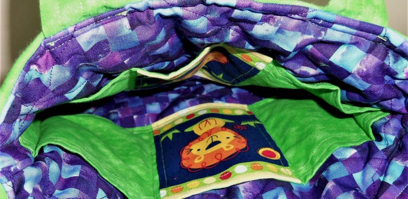 May include: A quilted fabric bag interior featuring a vibrant purple and blue checkered pattern lining. The bag includes bright green fabric pockets and a central pocket decorated with a cartoon lion graphic on a dark blue background with dots.