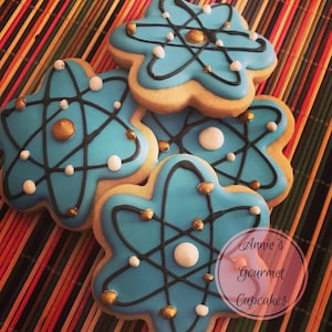 May include: Three blue and gold sugar cookies decorated with a black atom design. The cookies are stacked on top of each other. The cookies are on a brown and orange striped background. The text "Annie's Gourmet Cupcakes" is in a white circle on the bottom right of the image.
