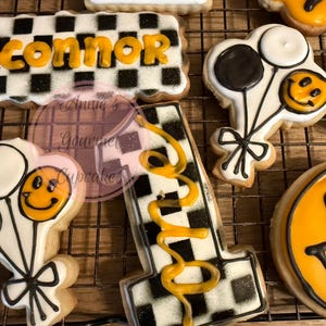 May include: Assortment of decorated sugar cookies, including a checkered rectangle with the name "Connor" in yellow, a number "1" with yellow icing, smiley face cookies, and balloon-shaped cookies. The cookies are on a wire rack.