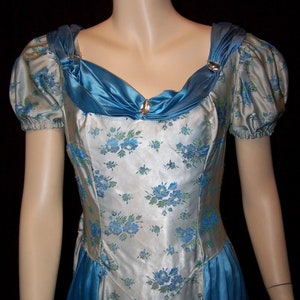 May include: A vintage-style dress with a floral pattern. The dress features a silver-white fabric with blue floral accents and a blue satin neckline and skirt panels. The sleeves are short and puffed, and the neckline is adorned with gold-colored clasps.