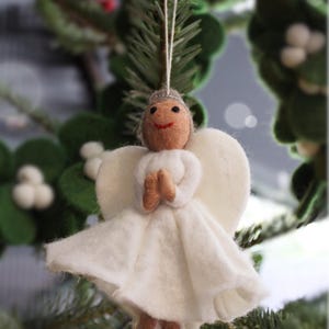 May include: A white felt angel ornament with a brown face and a white dress. The angel is hanging from a tree branch.