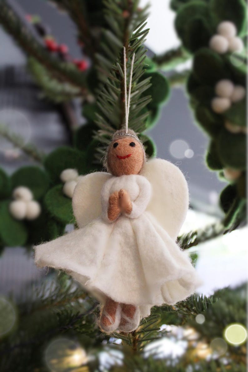 May include: A white felt angel ornament with a brown face and a white dress. The angel is hanging from a tree branch.