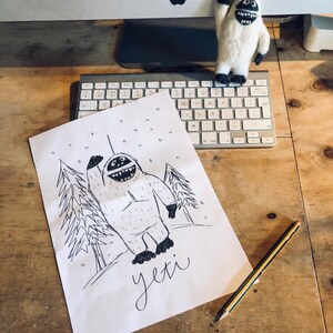 May include: A black and white drawing of a yeti waving with a snowy background and the word "yeti" written below. A small white yeti plush toy is sitting on a computer keyboard in the background.