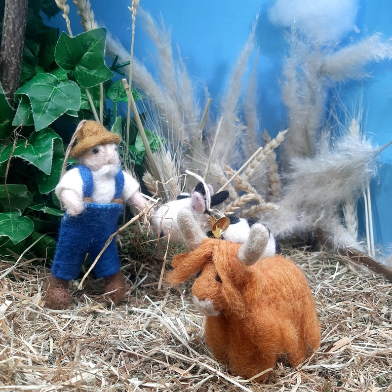 May include: A felt figurine of a farmer wearing a blue and white outfit and a straw hat, standing next to a brown and white cow and a brown highland cow figurine. The figurines are on a bed of straw.