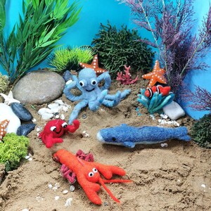 Op de afbeelding: Een blauwe vilt octopus, een rode vilt krab, een rode vilt kreeft, een blauwe vilt walvis en twee rode en witte vilt zeesterren op een zanderig oppervlak met groen mos en stenen.