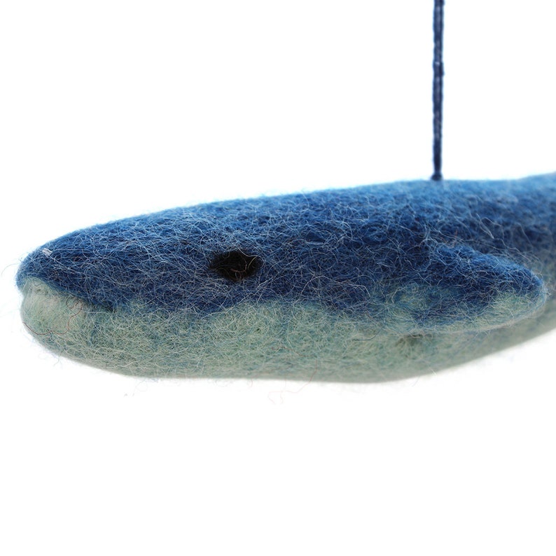 Op de afbeelding: Een blauwe vilten walvis-versiering die aan een blauwe touwtje hangt. De walvis heeft een witte buik en een zwart oog.