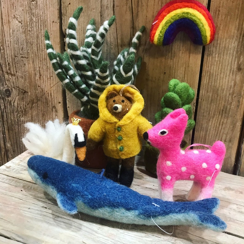 Op de afbeelding: Een set van vilt dieren ornamenten, waaronder een blauwe walvis, een roze hert, een gele beer met een regenjas en een witte zwaan. De ornamenten worden getoond op een houten oppervlak met een groene plant en een regenboog.