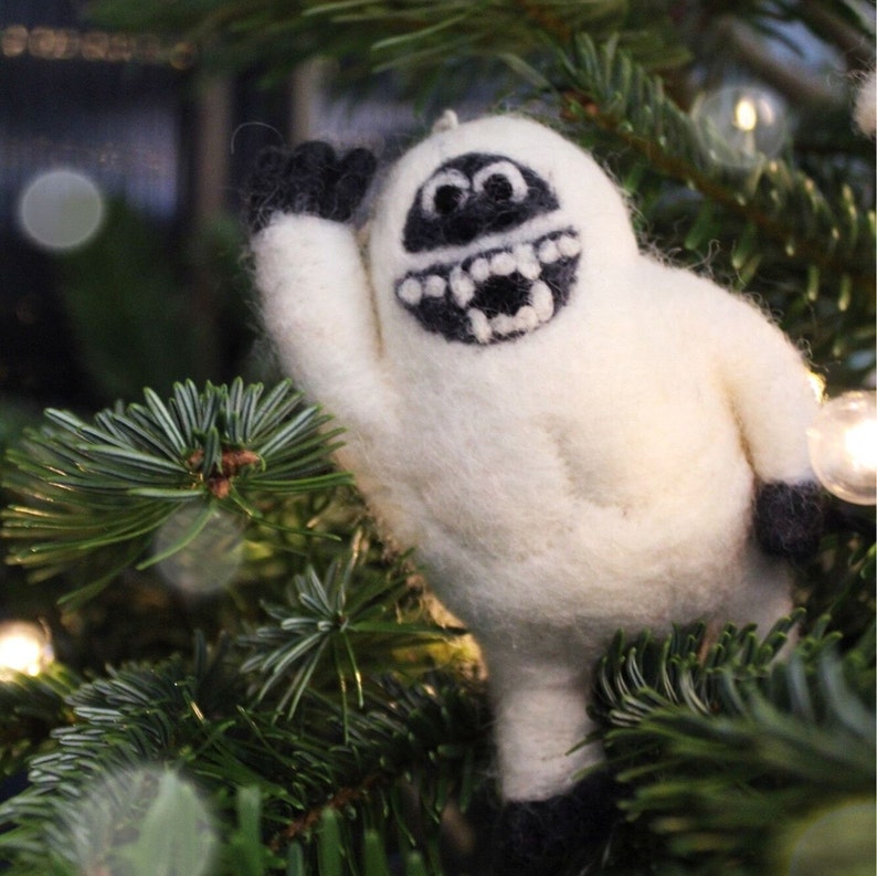 May include: A white felt snowman ornament with a black face and a wide, toothy grin. The snowman is waving with one arm raised.